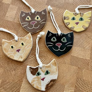 May include: Five ceramic cat-shaped ornaments with white rope hangers. The collection includes a solid brown, a yellow tabby, a light orange, a solid black, and a white, brown, and orange calico cat, each featuring painted eyes, noses, and whiskers.