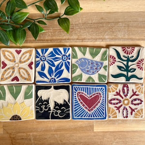 May include: Eight square ceramic tiles arranged in two rows. The designs include a yellow floral pattern, blue flowers, a blue bird on green, a red flower, a yellow sunflower, two black cats, a red heart with blue rays, and a red floral motif.