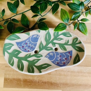 Op de afbeelding: Ovale keramische zeepbak met een handgeschilderd ontwerp met twee blauwe vogels te midden van groene bladeren. De schaal heeft twee afvoergaten en staat op een houten oppervlak met groen gebladerte.