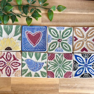 May include: A set of eight square ceramic tiles featuring hand-painted designs including a yellow sunflower, a red heart on a blue background, floral patterns in green, blue, and red, and a blue bird, all arranged in two rows on a wooden surface.