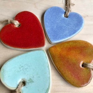 Op de afbeelding: Vier keramische harthornamenten in verschillende kleuren: rood, blauw, groen en oranje. Elk hart heeft een gat aan de bovenkant om op te hangen.