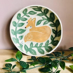 May include: A white ceramic bowl with a hand-painted design of a fox curled up in a bed of green leaves. The bowl is perfect for serving snacks or holding small items.