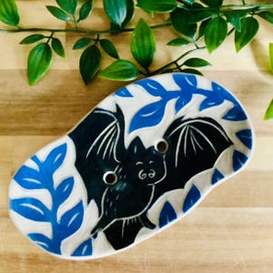 May include: A ceramic soap dish with a bat design. The dish is oval-shaped with a white base and blue leaf accents. The bat is black with white details and two holes for drainage.
