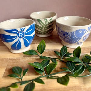 May include: Three small ceramic bowls with a cream-colored base. The left bowl features a large blue flower, the center bowl has green leaf patterns, and the right bowl displays a blue bird design with small heart accents and carved details.