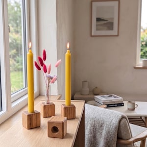 May include: Three square wooden blocks used as candle holders and a vase holder. Two blocks hold tall yellow taper candles with lit flames. One block holds a clear glass tube with pink and purple dried flowers. One empty block sits in front.