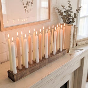 May include: A rectangular wooden candle holder base containing a row of eighteen lit taper candles in varying shades of white, cream, and light tan. The candles are arranged in a single line, each with a small flame, set within a dark wood block.