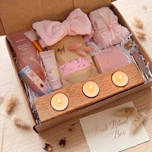 May include: A gift box containing a wooden candle holder with three lit tea lights, a pink plush headband, a pink towel with a ribbon, a Moika collagen wrapping mask, an Opos tube, a small box labeled AS, a thank you pouch, and a box of matches.