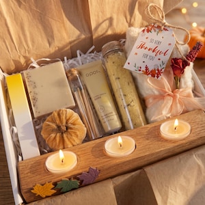 May include: A gift box containing a rectangular wooden candle holder with three lit tea lights, a bar of soap, a tube of lotion, a glass vial of matches, a glass tube of bath salts, and a white tag with the text So Very Thankful For You.