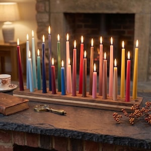 May include: A rectangular wooden candle holder with twenty-one holes, each containing a lit taper candle in various colors including red, blue, green, pink, yellow, and brown. The candles are arranged in a single row on a dark stone surface.