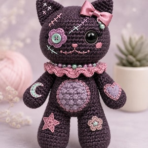May include: A dark purple crocheted cat doll featuring a pink satin bow, a ruffled collar with beads, and various stitched patches including a flower, a moon, a heart, and a star. The cat has one large black eye and one button eye.