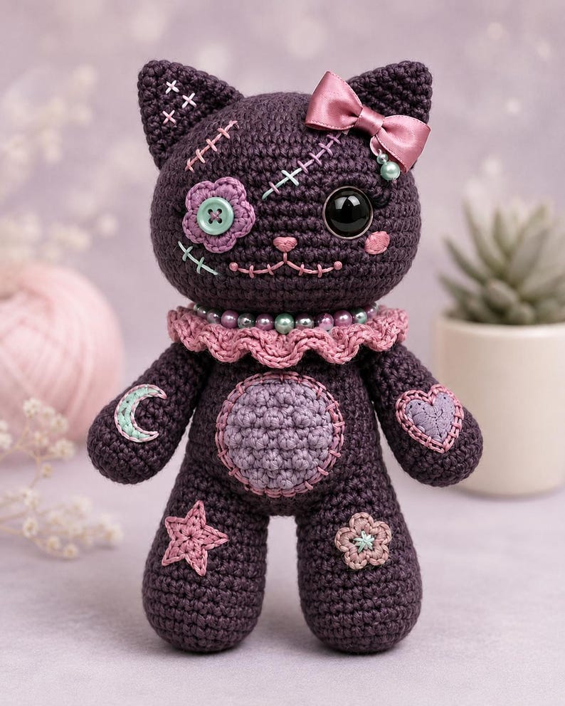 May include: A dark purple crocheted cat doll featuring a pink satin bow, a ruffled collar with beads, and various stitched patches including a flower, a moon, a heart, and a star. The cat has one large black eye and one button eye.