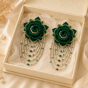 May include: A pair of dark green crocheted flower earrings featuring layered petals with gold-toned edges. Each flower is accented with a central cluster of small white pearls and hangs with multiple cascading strands of white pearls and green beads.
