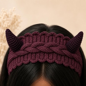 Puede incluir: Una diadema de punto de color morado oscuro con un patrón de trenza central, bordes festoneados y dos cuernos de ganchillo de color morado oscuro que sobresalen a cada lado. El accesorio está hecho de hilo texturizado de color ciruela.