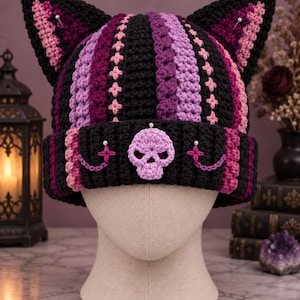 May include: A crocheted cat-ear beanie featuring vertical stripes in black, purple, and pink. The folded brim is adorned with a central light purple crocheted skull, two small silver-toned pins, and two delicate chains attached to the brim.