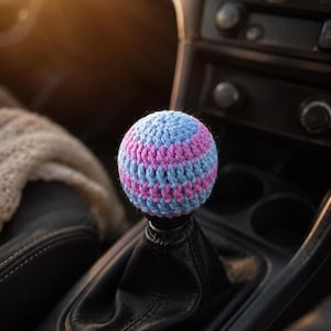 May include: A spherical crochet gear shift knob cover featuring alternating horizontal stripes of light blue and magenta yarn. The textured, handmade fabric cover is fitted snugly over a standard black automotive gear shifter handle.