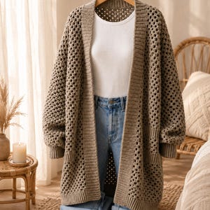 May include: A long-sleeved, open-front cardigan in a muted taupe colour, featuring a chunky crochet granny square pattern, ribbed cuffs, and a ribbed hem, layered over a plain white t-shirt and light blue denim jeans on a wooden hanger.