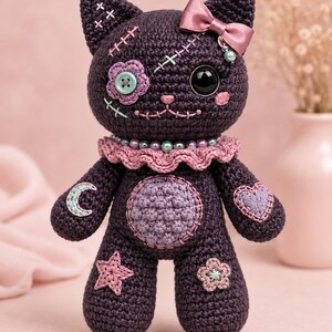 Puede incluir: Una muñeca de gato tejida a ganchillo en color morado oscuro con un lazo de satén rosa, un cuello con volantes y cuentas, y detalles cosidos. La muñeca tiene un ojo de botón, un parche de flor y formas bordadas de luna, estrella, corazón y flor.