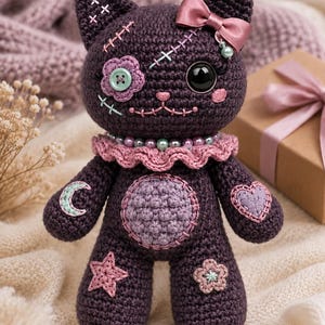 May include: A dark purple crocheted cat doll featuring a pink satin bow, a ruffled collar with beads, and stitched details. The doll has a button eye, a flower patch, and various embroidered shapes including a moon, heart, star, and flower.