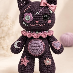 May include: A dark purple crocheted cat doll featuring a light purple belly patch, a pink ruffled collar with beads, and a satin bow. The doll has a button eye, a stitched flower eye, embroidered moon, star, and heart patches, and cross-stitch details.