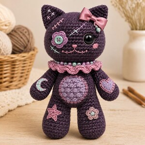 May include: A dark purple crocheted cat doll featuring a pink satin bow on one ear, a light blue button eye, a large black eye, and a ruffled pink collar. The body is decorated with embroidered patches including a crescent moon, a heart, a star, and a flower.