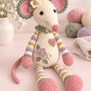 May include: A crocheted mouse toy with a cream-coloured body, pink ears, and striped arms and legs in shades of pink, yellow, green, and purple. The mouse features a small wooden button at the neck, a purple flower appliqué, and a pink heart patch.