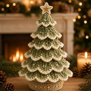Puede incluir: Un árbol de Navidad de ganchillo en verde salvia con bordes festoneados ribeteados en hilo crema. El árbol presenta pequeñas cuentas de perlas blancas y detalles de hilo dorado en todas las capas, rematado con una estrella de cinco puntas.