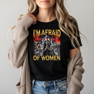 May include: A black short-sleeved t-shirt featuring a graphic print of three hooded skeletons with glowing red eyes, lightning bolts, and the text "I'M AFRAID OF WOMEN" in bold yellow letters. The shirt is worn under a beige knit cardigan.