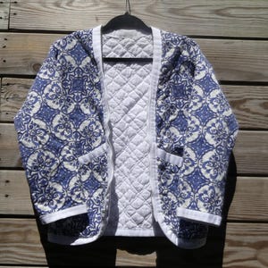 May include: A quilted jacket featuring a blue and white intricate floral pattern on the exterior and a solid white quilted interior. The garment has a front zipper closure, long sleeves, and white trim along the collar, cuffs, and hemline.