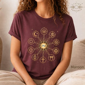 May include: A maroon short-sleeve t-shirt featuring a gold graphic design of various religious and spiritual symbols arranged in a circular pattern around the word ONE, which is centered in a sunburst motif on the front of the garment.