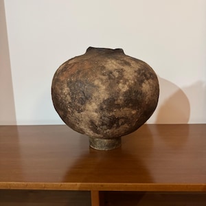 Pode incluir: Um vaso de cerâmica esférico com uma superfície texturizada em tons de castanho escuro e preto. A peça apresenta uma abertura circular estreita no topo e assenta numa pequena base cilíndrica, exibindo um aspeto terroso e envelhecido.