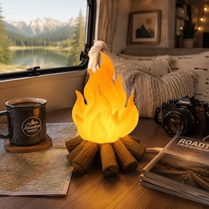 Könnte beinhalten: Eine dekorative Lagerfeuerlampe mit einem leuchtend orange-gelben, flammenförmigen Licht auf einem Sockel aus braunen, geformten Holzscheiten. Die Lampe steht auf einem Holztisch neben einer dunkelgrauen Tasse, einer Landkarte, einer Kamera und einem Buch.