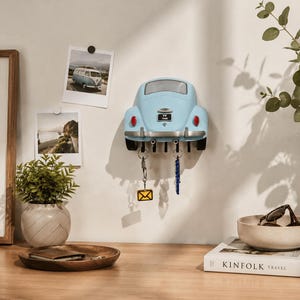 May include: A light blue wall-mounted key holder shaped like the rear of a vintage car. The design features a silver bumper, red taillights, and a black license plate with the text VW 1966. It includes five black hooks for hanging keys.