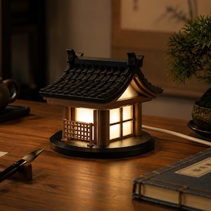 May include: A decorative lamp shaped like a traditional East Asian building with a dark tiled roof, light-coloured wooden walls, and a glowing interior. The structure features a small lattice fence detail and sits on a circular black base with a power cord.