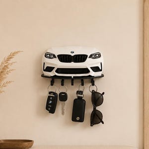 May include: A white wall-mounted key holder shaped like the front end of a BMW car, featuring a black grille, headlights, and five black hooks. The hooks hold various items including a car key fob, a metal key, a black key case, and sunglasses.