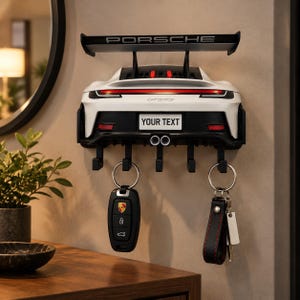 Könnte beinhalten: Ein Wand-Schlüsselhalter in Form des Hecks eines weißen Porsche GT3RS Sportwagens. Er verfügt über einen großen schwarzen Heckspoiler mit dem Schriftzug PORSCHE, ein Nummernschildfeld mit der Aufschrift YOUR TEXT, zwei Auspuffrohre und fünf schwarze Haken.