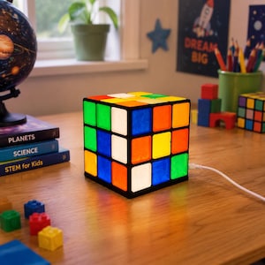 Könnte beinhalten: Eine quadratische Lampe, die wie ein klassischer, mehrfarbiger Puzzlewürfel gestaltet ist. Die Lampe hat einen schwarzen Rahmen mit beleuchteten, durchscheinenden Feldern in Rot, Blau, Grün, Gelb, Orange und Weiß. Ein weißes Stromkabel ragt heraus.