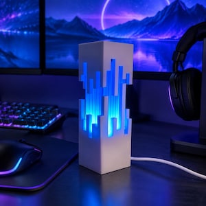 Puede incluir: Una lámpara de torre rectangular blanca con un patrón de corte vertical que emite una luz azul brillante desde el interior. La lámpara tiene un acabado mate suave y está conectada a un cable de alimentación blanco, colocada sobre una superficie.