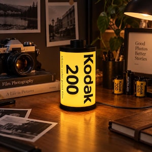 Puede incluir: Una lámpara cilíndrica diseñada para parecerse a un rollo de película Kodak 200 35mm color print film. La lámpara presenta un cuerpo amarillo con texto negro que dice Kodak 200 y 35mm color print film, emitiendo una luz cálida interna.