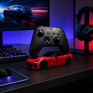 May include: A black wireless game controller resting on top of a red sports car model. The controller features a central Xbox logo, a directional pad, two analog sticks, and four colored action buttons labeled A, B, X, and Y. The car model is red.
