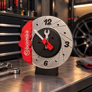 Puede incluir: Un reloj de pared circular diseñado para parecer un disco de freno perforado plateado con una pinza de freno roja Brembo en el lado izquierdo. La esfera presenta los números negros 12, 3 y 6, con un destornillador y una llave como manecillas.
