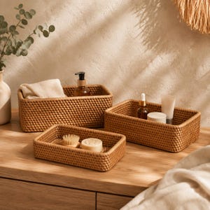 Peut inclure: Trois paniers de rangement rectangulaires en rotin tressé de couleur brun clair. Le plus grand contient un tissu beige plié et un flacon à pompe brun. Le moyen contient un flacon compte-gouttes en verre et un tube. Le plus petit contient une brosse et un savon.