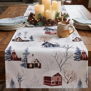 May include: A white table runner featuring a repeating winter pattern of snow-covered cabins, evergreen trees, and bare branches. The runner is placed on a wooden table with stacks of light blue napkins, a ceramic mug, and a candle centerpiece.