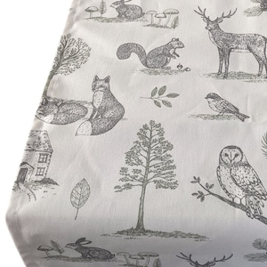 Op de afbeelding: Een witte stof met een bosmotief met verschillende dieren, waaronder een vos, een eekhoorn, een uil, een hert, een konijn en vogels. Het patroon is in grijstinten en bruin.