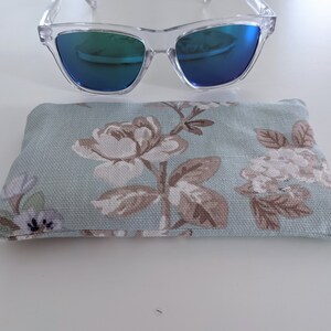 Può includere: Occhiali da sole con montatura trasparente e lenti a specchio blu sopra un astuccio per occhiali con motivo floreale. L'astuccio ha uno sfondo azzurro con un motivo ripetuto di fiori e foglie bianche e marroni.