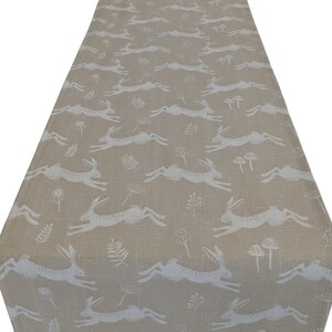 Può includere: Runner da tavolo beige con un motivo ripetuto di lepri bianche in corsa, funghi ed elementi floreali. Il tessuto ha un aspetto strutturato, adatto per un tavolo da pranzo o un'esposizione decorativa. Il runner è di colore neutro.
