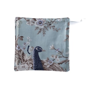 May include: Square, light blue fabric item with a peacock and floral design. The peacock is dark blue with a detailed head and neck. The flowers are white, light blue, and brown. A white loop is attached to the top corner.
