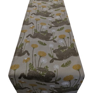 Op de afbeelding: Een beige tafelkleed met een herhalend patroon van bruine konijnen die door een veld met gele en witte paardenbloemen rennen.