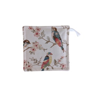 Puede incluir: Un posavasos de tela beige con un patrón floral con pájaros coloridos y flores rosas. El posavasos tiene un asa de bucle blanco.