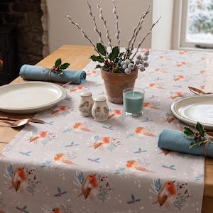 May include: A light gray table runner featuring a repeating pattern of robins, holly leaves, and berries. The runner is set on a wooden table with two white plates, two blue cloth napkins, copper-colored cutlery, salt and pepper shakers, and a candle.