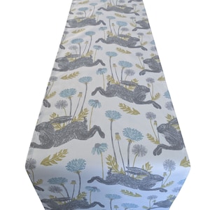 Può includere: Un runner da tavolo bianco con un motivo ripetuto di conigli grigi che corrono attraverso un campo di dente di leone blu e verdi.